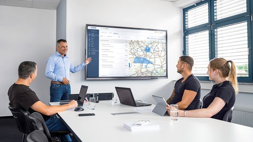 A fleet manager lectures a team of two men and one woman in a bright, modern meeting room. He points to a large screen and his team listens attentively.